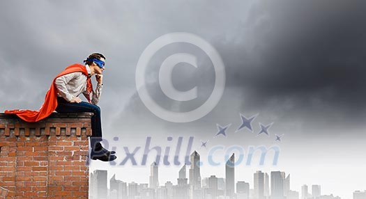 Thoughtful superman in cape and mask sitting on top of building Thoughtful superman in cape and mask sitting on top of building