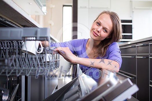 Pretty, young woman in her modern and well equiped kitchen putting cups into the dishwasher - an appliance that helps her keep the home tidy Pretty, young woman in her modern and well equiped kitchen putting cups into the dishwasher - an appliance that helps her keep the home tidy