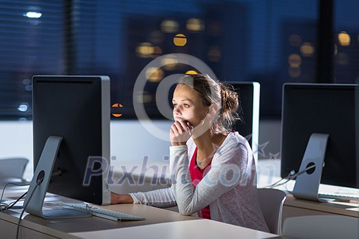Pretty, young female college student using a desktop computer/pc in a college library - burning the midnight oil, working hard toward her goals instead of getting enough sleep (shallow DOF; color toned image)