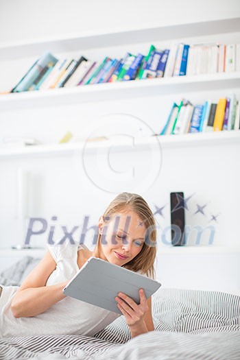 Pretty, young woman lying in bed, using her tablet computer, reading sending e-mails, using apps, having fun Pretty, young woman lying in bed, using her tablet computer, reading sending e-mails, using apps, having fun