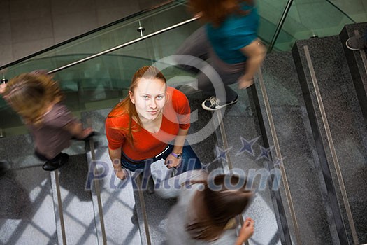 At the university/college - Students rushing up and down a busy stairway - confident pretty young female student looking upwards (color toned image) At the university/college - Students rushing up and down a busy stairway - confident pretty young female student looking upwards (color toned image)