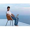 handsome young man relaxing and working on laptop computer at home balcony while looking sunset