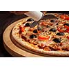Pizza cutter wheel slicing ham pizza with capsicum and olives on wooden board on table