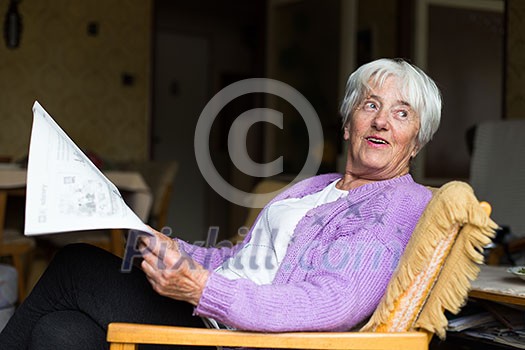 Senior woman reading morning newspaper, sitting in her favorite chair in her living room, looking happy Senior woman reading morning newspaper, sitting in her favorite chair in her living room, looking happy