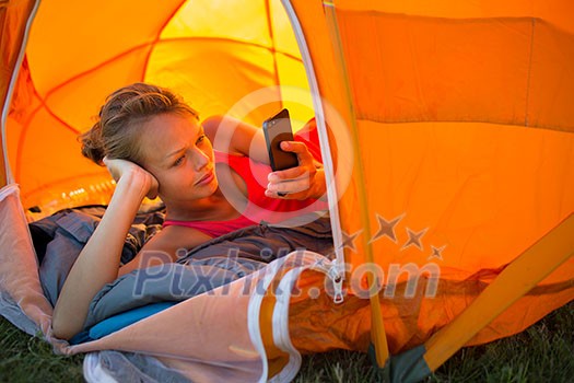 Pretty, young woman camping outdoors, lying in the tent in the morning, using her smart phone Pretty, young woman camping outdoors, lying in the tent in the morning, using her smart phone