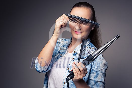 Funny portrait of a young female hairstylist, holding her tools and posing with a comb in front of her eyes ion front of a grey backdrop (color toned image; shallow DOF) Funny portrait of a young female hairstylist, holding her tools and posing with a comb in front of her eyes ion front of a grey backdrop (color toned image; shallow DOF)