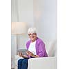 Portrait of a senior woman at home using a tablet computer - Looking happy, looking at the camera, smiling while sitting on the sofa in her living room