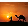 Rajasthan travel background - indian cameleer (camel driver) with camel silhouette in dunes of Thar desert on sunset. Jaisalmer, Rajasthan, India