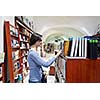 Pretty female student standing at bookshelf in university library store shop  searching for a book