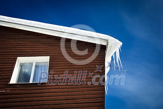 Winter/mountain concept: closeup of a wooden cottage roof covered with snow and icicles against blue sky Winter/mountain concept: closeup of a wooden cottage roof covered with snow and icicles against blue sky