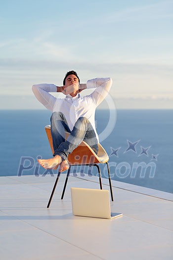 handsome young man relaxing and working on laptop computer at home balcony while looking sunset