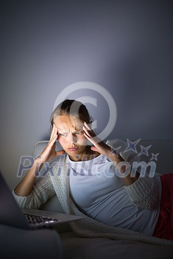 Very tired young woman, burning the midnigh oil - working late at night on her laptop computer, at home, sitting on sofa, rubbing her tired eyes, trying to stay focused