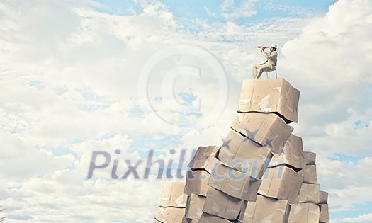 Businessman sitting on big stack of carton boxes and looking in spyglass Businessman sitting on big stack of carton boxes and looking in spyglass