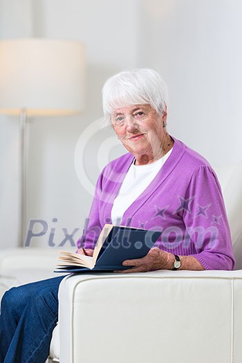Portrait of a senior woman at home - Looking happy, looking at the camera, smiling while sitting on the sofa in her living room and reading a good book Portrait of a senior woman at home - Looking happy, looking at the camera, smiling while sitting on the sofa in her living room and reading a good book