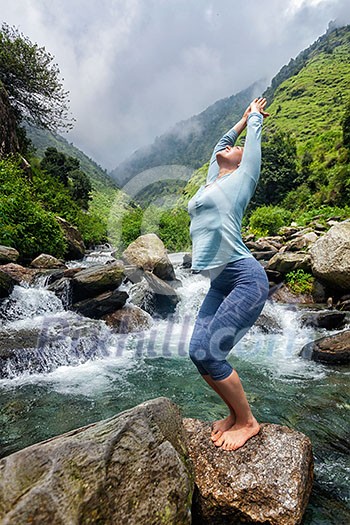 Young sporty fit woman doing yoga asana Utkatasana (chair pose) outdoors at tropical waterfall standing on stone Young sporty fit woman doing yoga asana Utkatasana (chair pose) outdoors at tropical waterfall standing on stone