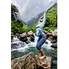 Young sporty fit woman doing yoga asana Utkatasana (chair pose) outdoors at tropical waterfall standing on stone