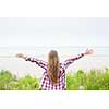 Back view of young woman with long blond hair and outstretched arms on empty beach facing ocean wearing plaid shirt