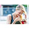 Beautiful young blonde woman applying lipstick with compact makeup mirror outside on city street