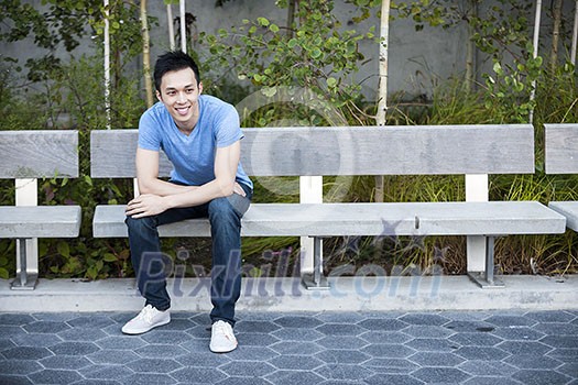 Young asian man sitting and smiling on park bench with copy space Young asian man sitting and smiling on park bench with copy space