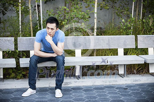 Concerned young asian man sitting on park bench with copy space Concerned young asian man sitting on park bench with copy space
