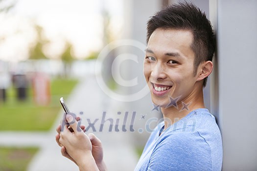 Smiling young asian man holding smart phone and looking at camera on city sidewalk Smiling young asian man holding smart phone and looking at camera on city sidewalk