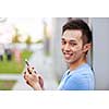 Smiling young asian man holding smart phone and looking at camera on city sidewalk