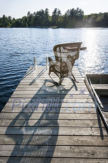 Wicker rocking chair on wooden dock in summer at small lake casting long shadow Wicker rocking chair on wooden dock in summer at small lake casting long shadow