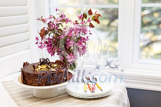 Round gourmet chocolate cake on table next to window with plates, cutlery, and pink spring bouquet Round gourmet chocolate cake on table next to window with plates, cutlery, and pink spring bouquet
