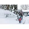Man using snowblower to clear deep snow on driveway near residential house after heavy snowfall.