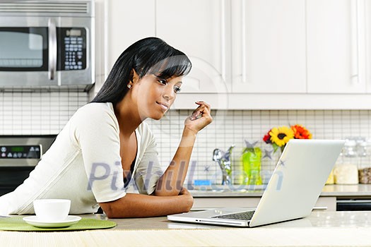 Thoughtful black woman using computer in modern kitchen interior Thoughtful black woman using computer in modern kitchen interior