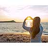 Blonde young girl holding hands in heart shape framing setting sun at sunset on ocean beach