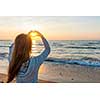 Blonde young girl holding hands in heart shape framing setting sun at sunset on ocean beach