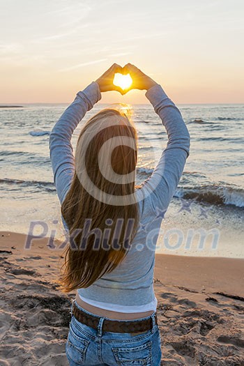 Blonde young girl holding hands in heart shape framing setting sun at sunset on ocean beach Blonde young girl holding hands in heart shape framing setting sun at sunset on ocean beach
