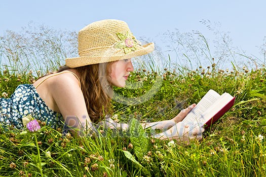 Young teenage girl reading book in summer meadow with straw hat