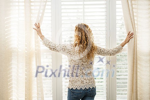Woman looking out big bright window with curtains and blinds Woman looking out big bright window with curtains and blinds