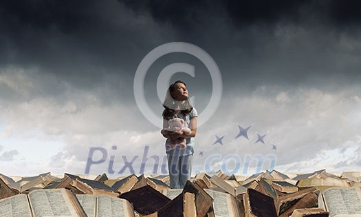Cute girl wearing pajamas with toy bear in hand standing on pile of books Cute girl wearing pajamas with toy bear in hand standing on pile of books