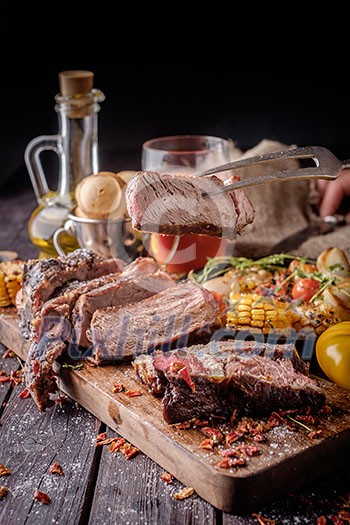 Piece grilled steak on a fork. Grilled steak sliced on a cutting board. Entrecote with vegetables on a wooden background. Piece grilled steak on a fork. Grilled steak sliced on a cutting board. Entrecote with vegetables on a wooden background.
