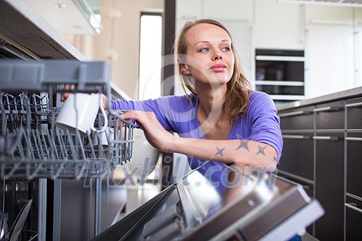 Pretty, young woman in her modern and well equiped kitchen putting cups into the dishwasher - an appliance that helps her keep the home tidy Pretty, young woman in her modern and well equiped kitchen putting cups into the dishwasher - an appliance that helps her keep the home tidy