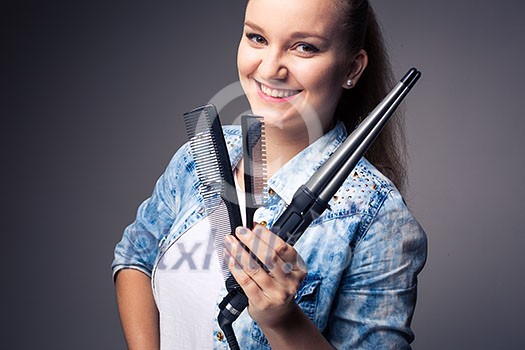 Funny portrait of a young female hairstylist, holding her tools and posing with a comb in front of her eyes ion front of a grey backdrop (color toned image; shallow DOF) Funny portrait of a young female hairstylist, holding her tools and posing with a comb in front of her eyes ion front of a grey backdrop (color toned image; shallow DOF)