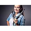 Funny portrait of a young female hairstylist, holding her tools and posing with a comb in front of her eyes ion front of a grey backdrop (color toned image; shallow DOF)