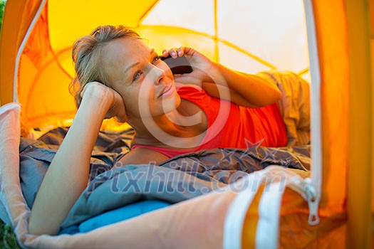 Pretty, young woman camping outdoors, lying in the tent in the morning, using her smart phone Pretty, young woman camping outdoors, lying in the tent in the morning, using her smart phone