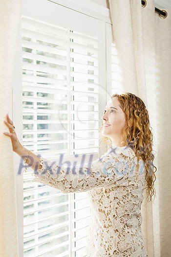 Happy woman looking out big bright window with curtains and blinds Happy woman looking out big bright window with curtains and blinds