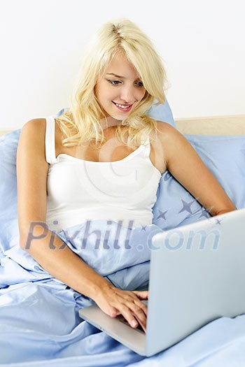 Happy blonde woman using computer at home sitting in bed