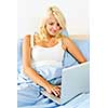 Happy blonde woman using computer at home sitting in bed