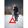 Young woman setting up a warning triangle and calling for assistance after her car broke down in the middle of nowhere on a freezing winter day