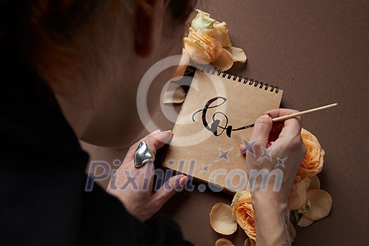 Women's Day. Notebook used for ideas and positive emotions. Yellow roses around it. Woman's hands writing word love in diary or notebook. Women's Day. Notebook used for ideas and positive emotions. Yellow roses around it. Woman's hands writing word love in diary or notebook.