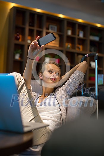 Young female passenger at the airport, about to check-in, enjoying the waiting time in the lounge, taking selfies with her smartphone (color toned image; shallow DOF)