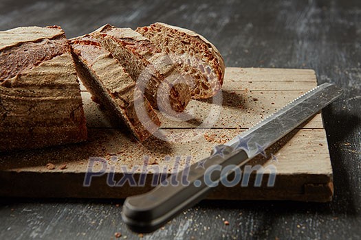 Loaf of sliced whole grain bread on wood bread board with knife, on a dark background