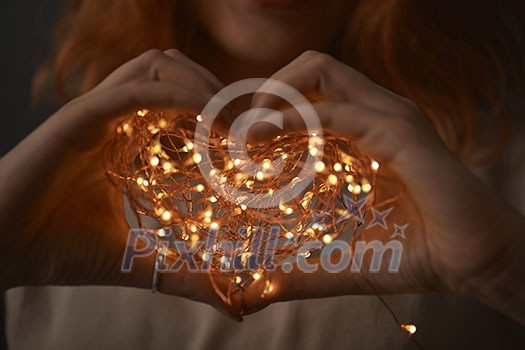 Female hands holding a Christmas string of lights in the form of a heart on a dark background Female hands holding a Christmas string of lights in the form of a heart on a dark background