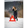 Young woman setting up a warning triangle and calling for assistance after her car broke down in the middle of nowhere on a freezing winter day